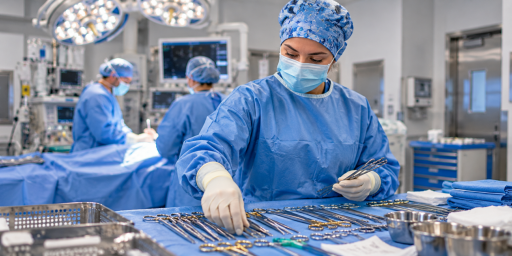 A masked surgical technologist wearing a blue gown and gloves arranges surgical instruments on a sterile draped table in an operating room.