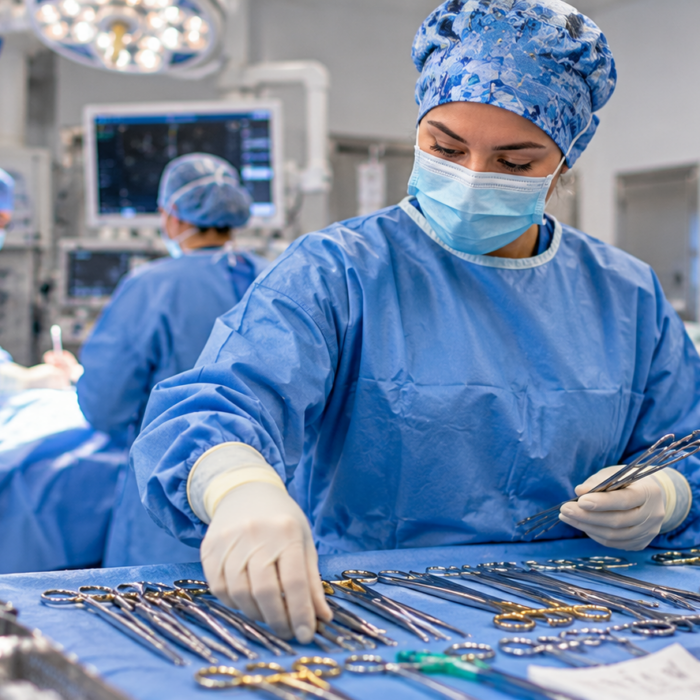 A masked surgical technologist wearing a blue gown and gloves arranges surgical instruments on a sterile draped table in an operating room.