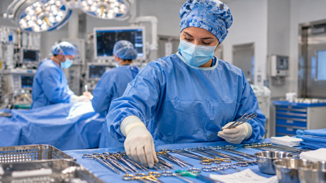 A masked surgical technologist wearing a blue gown and gloves arranges surgical instruments on a sterile draped table in an operating room.